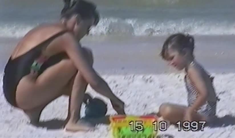 A nostalgic home video from 1997 shows a mother and her young daughter playing together in the sand on a sunny beach by the ocean