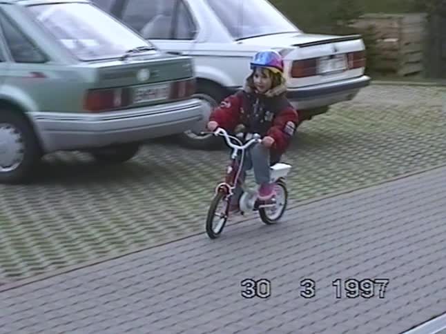 A little girl wearing a bright helmet proudly rides her small bicycle around a paved parking lot in this charming vintage home video from 1997
