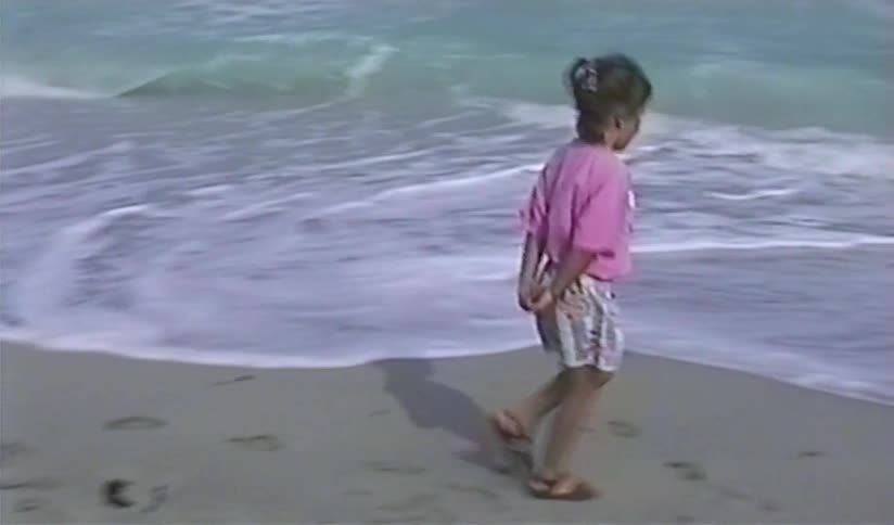 A young girl in a pink shirt happily walks along the shoreline as waves crash on the sandy beach in this vintage home movie footage