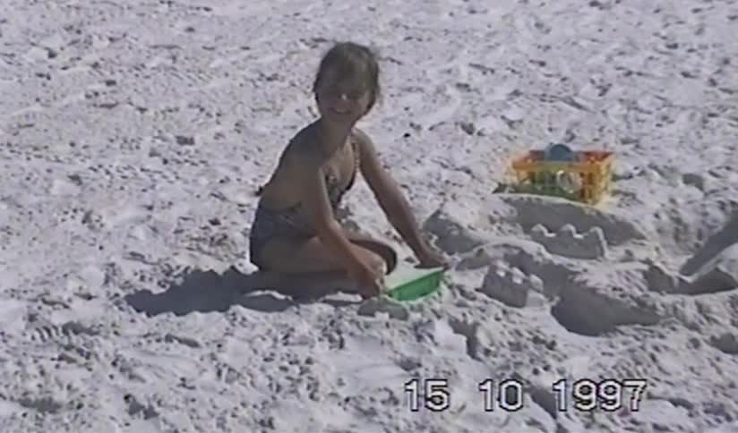 A young girl smiles while building a sandcastle on a bright, sunny beach in this nostalgic home video from 1997