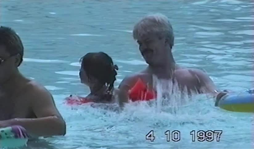 A father with a mustache holds his young daughter with floaties as she happily splashes in a swimming pool in a nostalgic home video from 1997