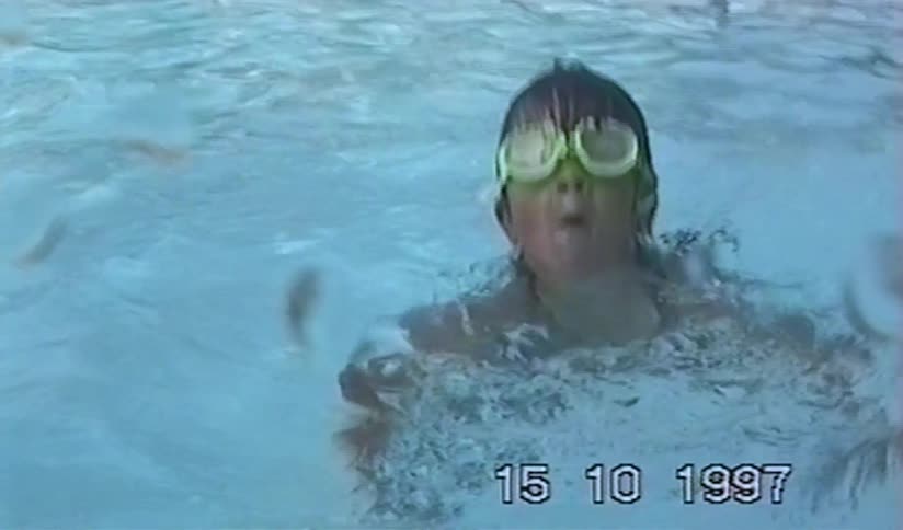 A young girl with bright green goggles splashes around in a swimming pool in a nostalgic home video from 1997