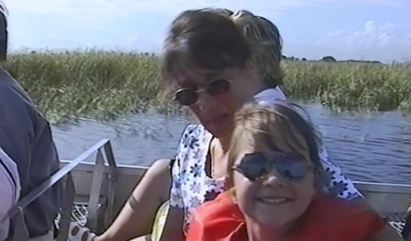A vintage home video shows a smiling mother and daughter enjoying an airboat ride through a sunny marsh