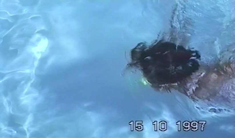 A vintage home movie from 1997 shows a young girl with bright green goggles swimming and smiling in a pool