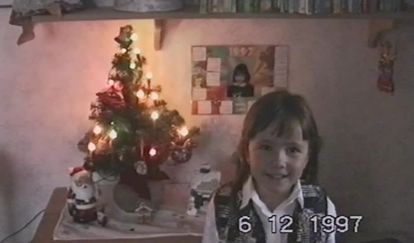 A vintage home video from 1997 shows a cute young girl smiling next to a small, decorated Christmas tree