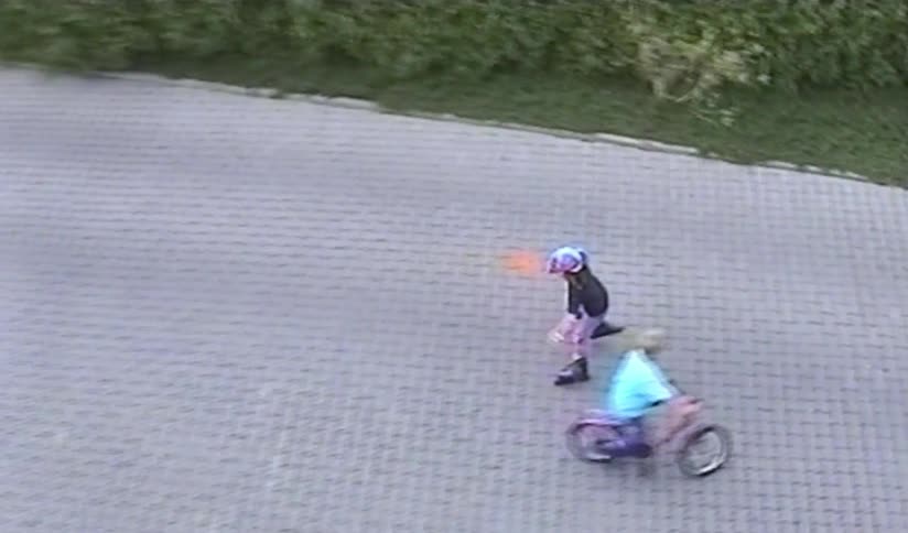 A nostalgic home video from a high-angle perspective shows a young girl in a colorful helmet and pads rollerblading on a paved driveway