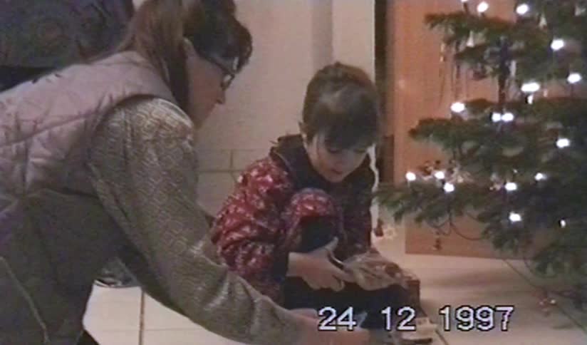A mother helps her young daughter open Christmas presents next to the tree in a nostalgic home video from Christmas Eve 1997