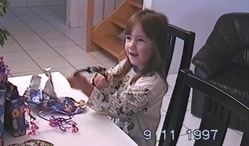 A cute little girl in pajamas happily unwraps a birthday present at the table in a home video from 1997