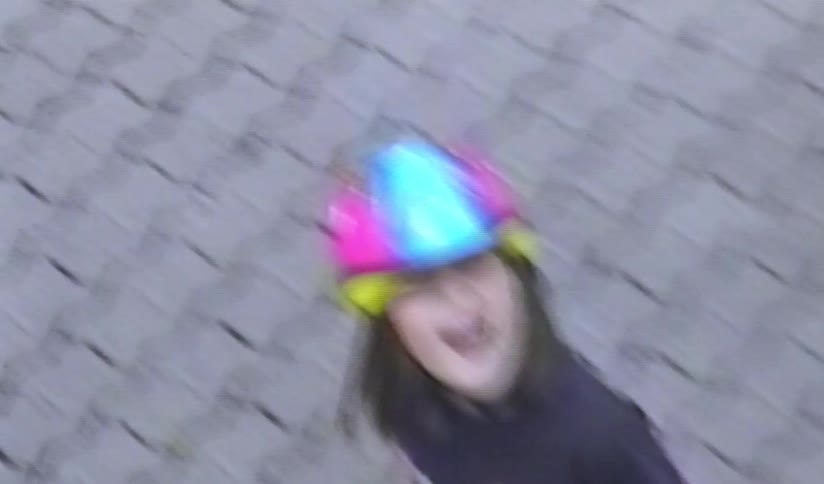A high-angle shot from a vintage home video shows a little girl in a colorful helmet rollerblading on a patio before stopping to smile at the camera