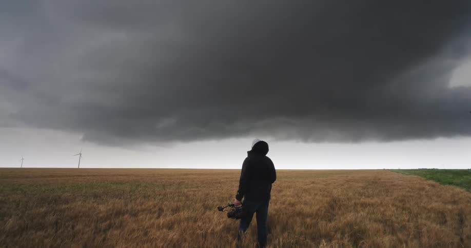 A storm chaser holding a camera walks into a vast wheat field to film a massive, dark supercell storm cloud looming on the horizon