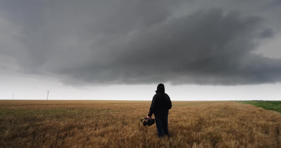 A filmmaker with a camera stands alone in a vast golden wheat field, facing an ominous and dramatic approaching storm cloud