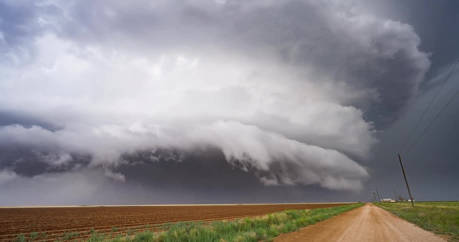 A massive supercell thunderstorm with a dramatic shelf cloud looms over a rural dirt road and farm fields