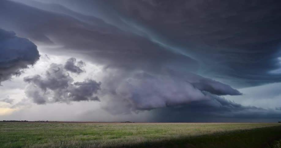 A dramatic and imposing supercell thunderstorm with dark, swirling clouds looms over a vast, green prairie