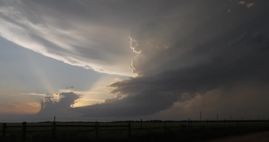 Massive storm clouds are illuminated by the setting sun, casting dramatic rays over a vast green field