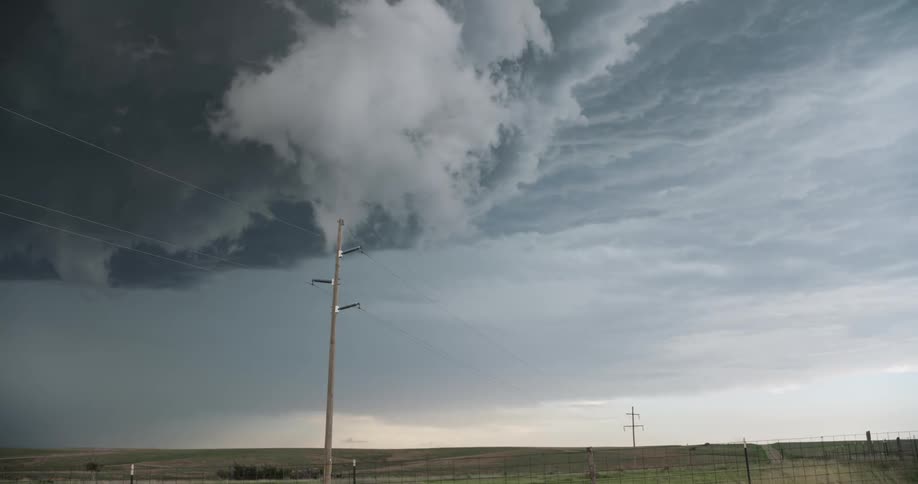 A massive, dark supercell storm cloud hovers ominously over a rural landscape with power lines in the foreground