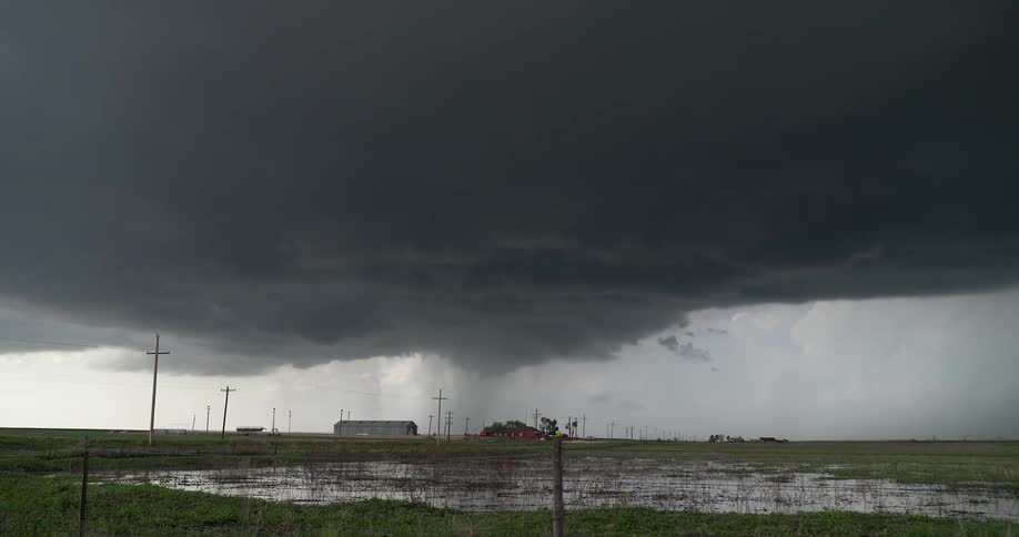 An ominous supercell thunderstorm hovers over a flooded rural field, dropping a heavy column of rain