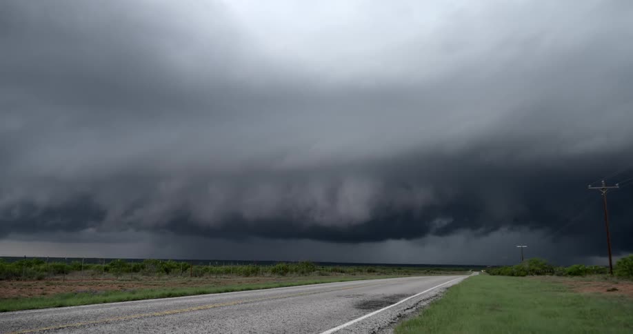 A massive, dark shelf cloud from a severe thunderstorm looms ominously over an empty rural road