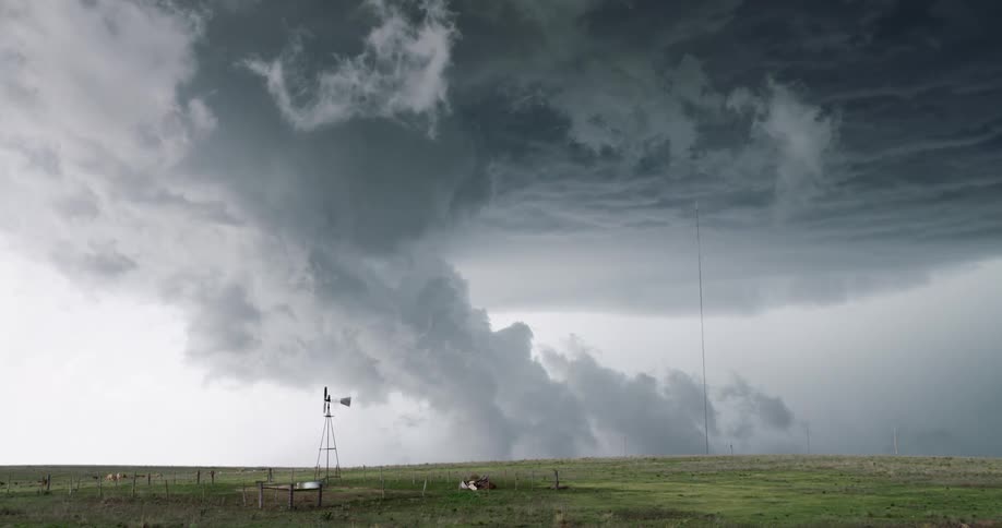 An incredibly powerful supercell thunderstorm forms a dangerous tornado over a peaceful rural farm with a windmill