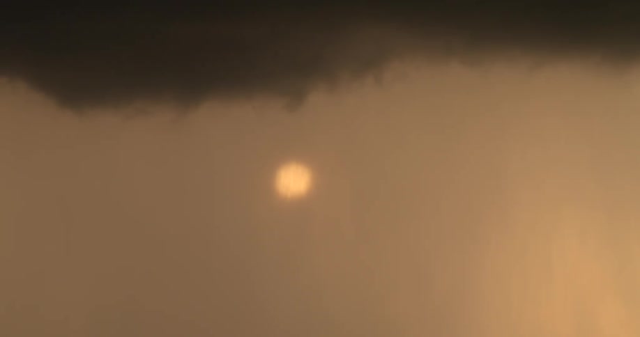 A telephoto shot captures the sun glowing brightly through a hazy, orange sky beneath a dark cloud bank