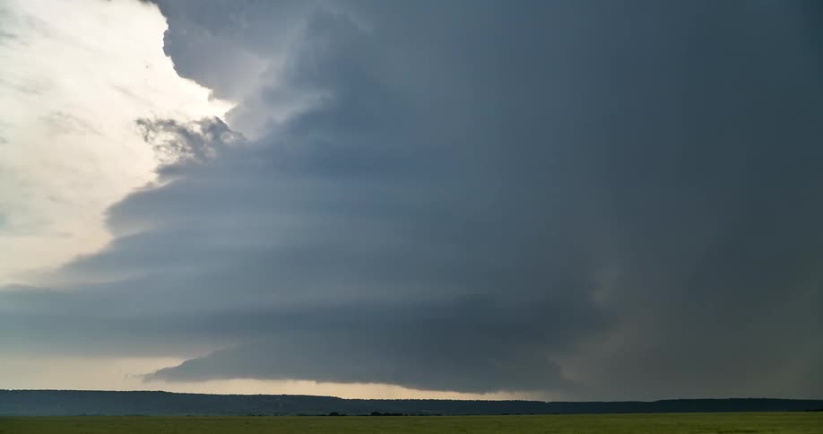 A massive supercell thunderstorm forms over a vast green field, its dark, swirling clouds signaling an approaching storm