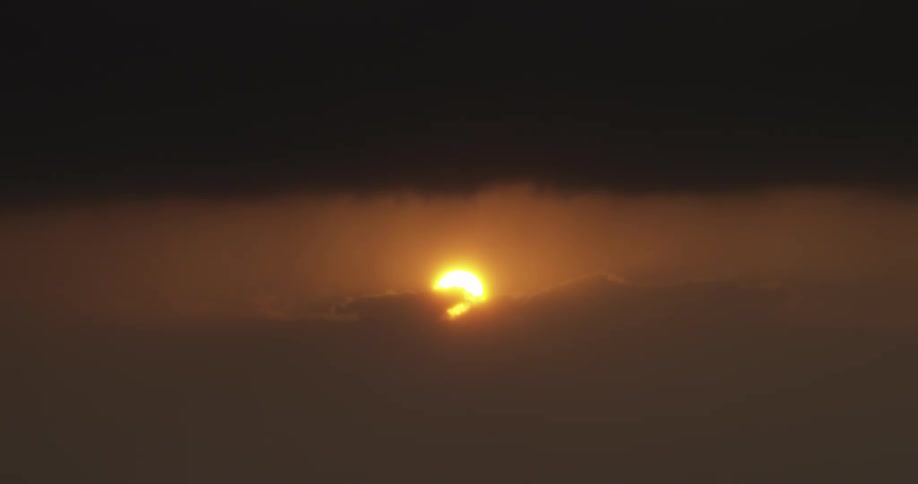 The warm golden sun peeks over a bed of dark, hazy clouds at sunset
