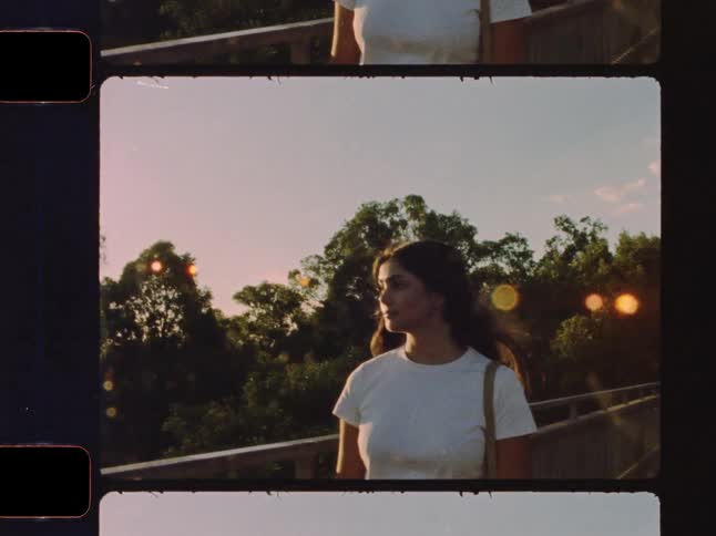 Dreamy, vintage-style footage captures a young woman standing on a wooden bridge in a park at golden hour