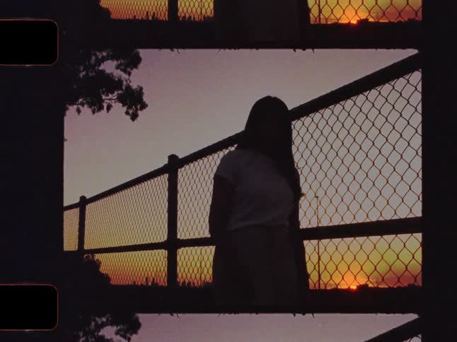 Vintage Super 8 film captures a silhouetted young woman watching the sunset from behind a chain-link fence on a bridge