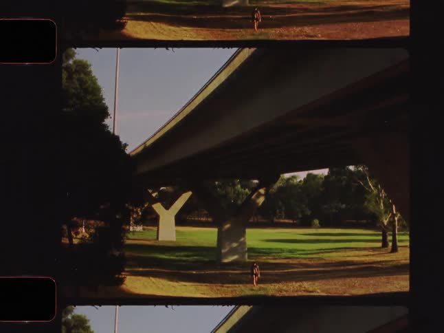A solitary person walks under a modern concrete bridge in a sunny park on a quiet afternoon, captured on vintage film