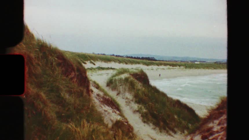A vintage film shot from grassy sand dunes captures a wide view of a person walking alone on a deserted beach on a cloudy day