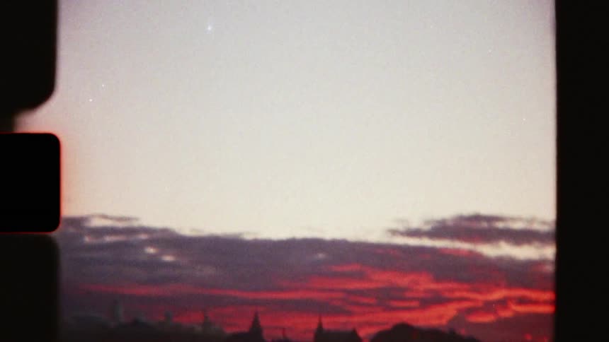 Vintage 8mm footage of a bird soaring in the sky, revealing a blurry city skyline against a beautiful red sunset