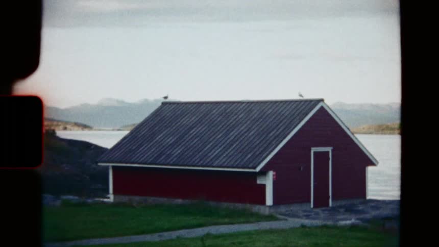 A slow zoom on a red boathouse by a calm lake in the countryside, shot on vintage 8mm film