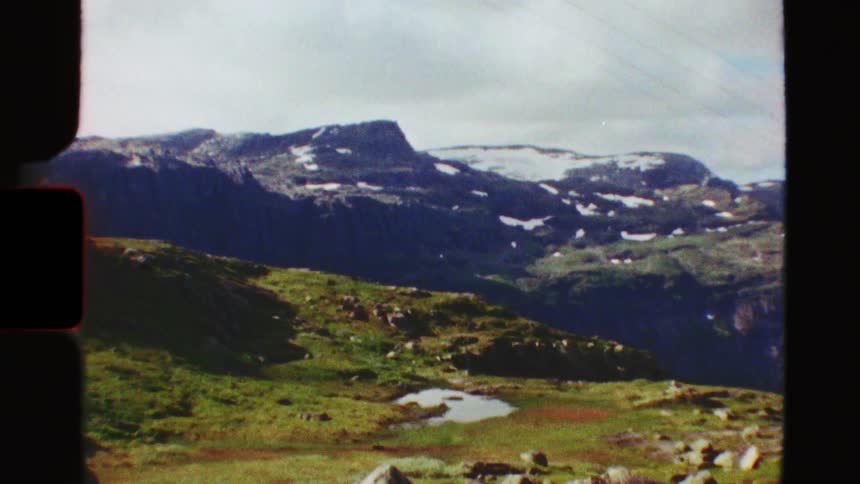 Vintage 8mm film captures a serene view of snow-capped mountains and a lush green valley with a small pond