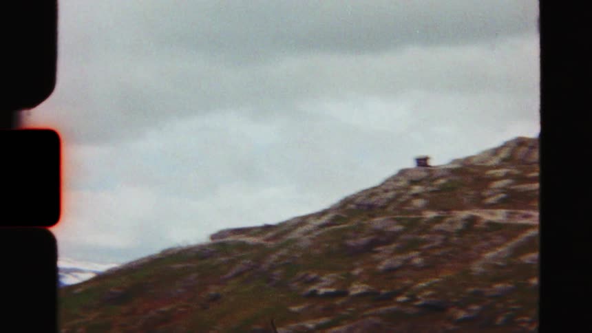 Vintage film captures hikers ascending a rocky mountain trail towards a lone hut at the peak on a cloudy day