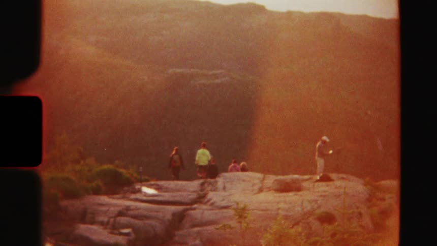 Vintage 8mm footage shows a photographer and friends on a rocky mountain overlook enjoying a golden hour sunset