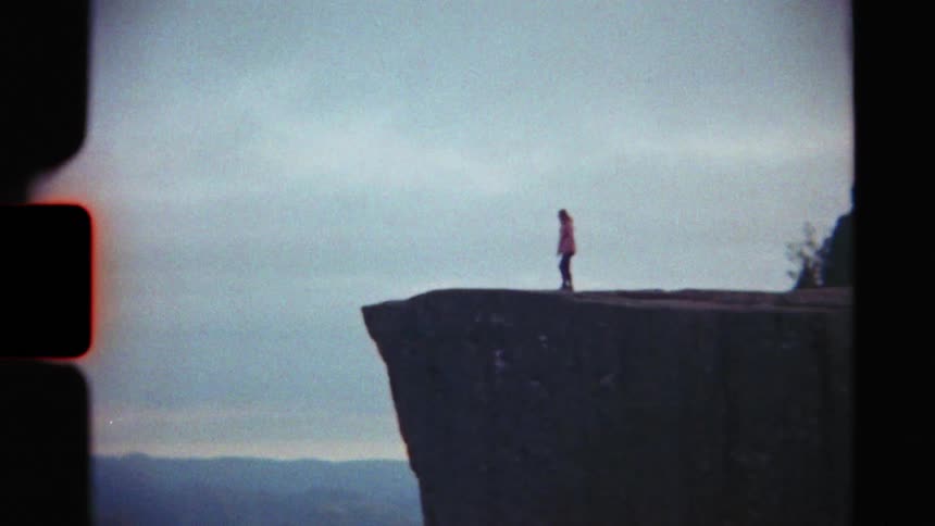 A vintage 8mm film shot shows a solitary person in a pink jacket walking along the edge of a dramatic, flat-topped cliff overlooking a vast valley