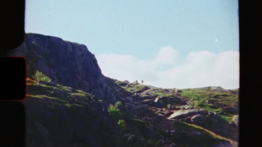 Super 8 film captures distant hikers making their way up a winding path on a green and rocky mountainside
