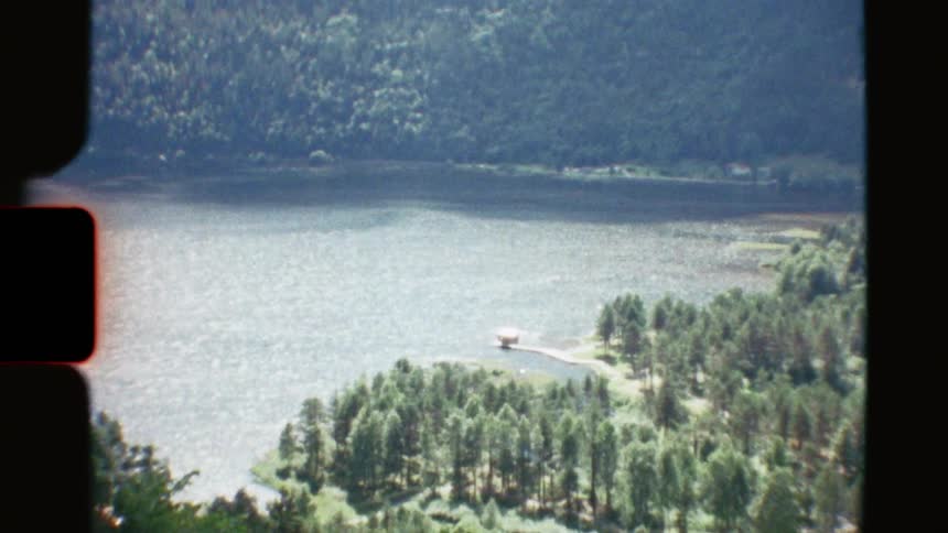 An archival high-angle shot of a secluded boathouse on the shore of a vast, shimmering lake