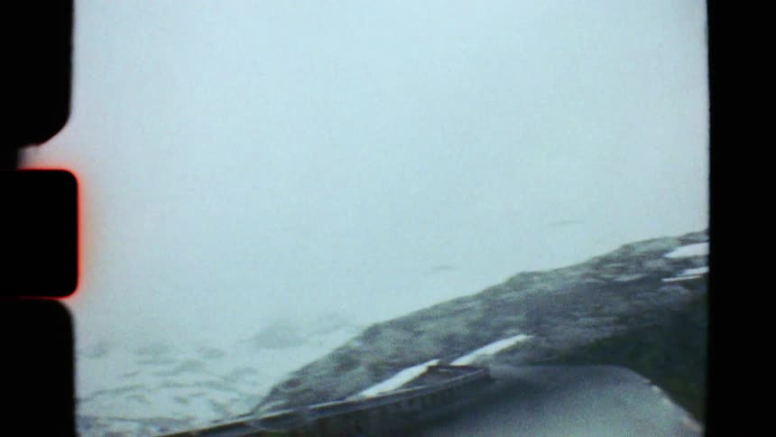 Authentic Super 8 footage captures a car driving through a foggy and rainy mountain pass with lingering snow