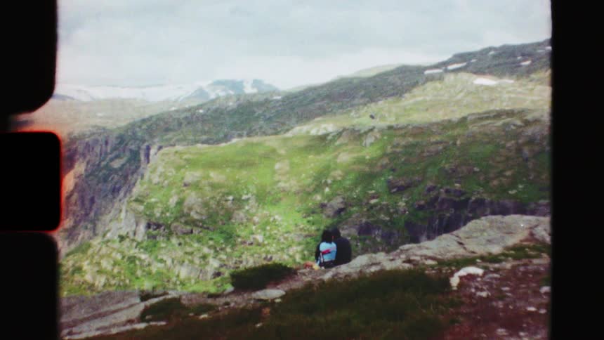 Vintage Super 8 footage zooms out from a couple sitting on a cliff's edge to reveal a breathtaking mountain landscape