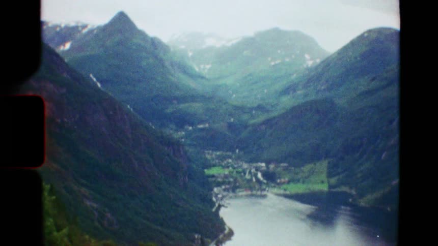 Vintage Super 8 footage pans up from a car on a winding cliffside road to reveal the majestic Geiranger Fjord in Norway