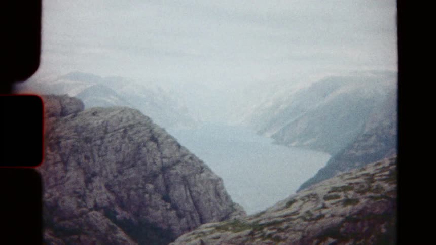Vintage 8mm film captures hikers on a rocky mountain trail revealing a stunning view of a distant fjord