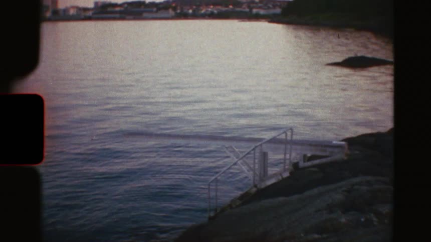 A vintage film reveals a quiet coastal town at dusk, focusing on a lone diving board over the calm, rippling water