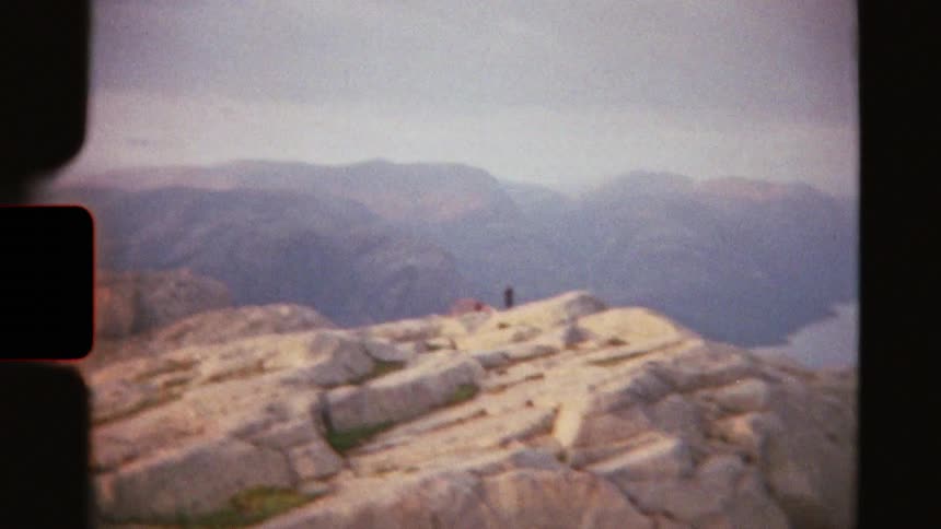 Vintage film captures hikers standing on a vast, rocky mountain peak overlooking a winding fjord below