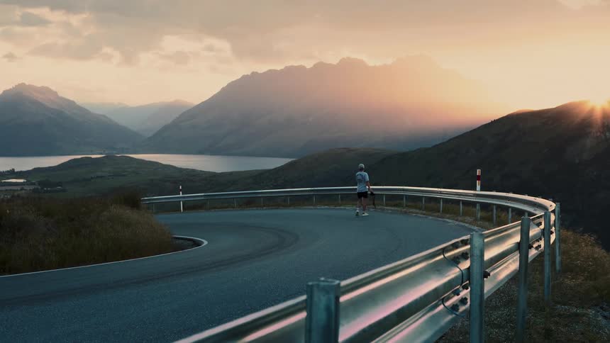 A man jogs on a winding mountain road carrying a tripod and camera with a stunning sunset over a lake in the background