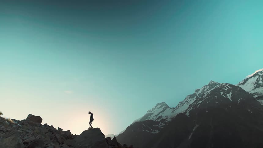 A hiker is silhouetted against the rising sun as they trek across a rocky mountain ridge with snowy peaks in the distance