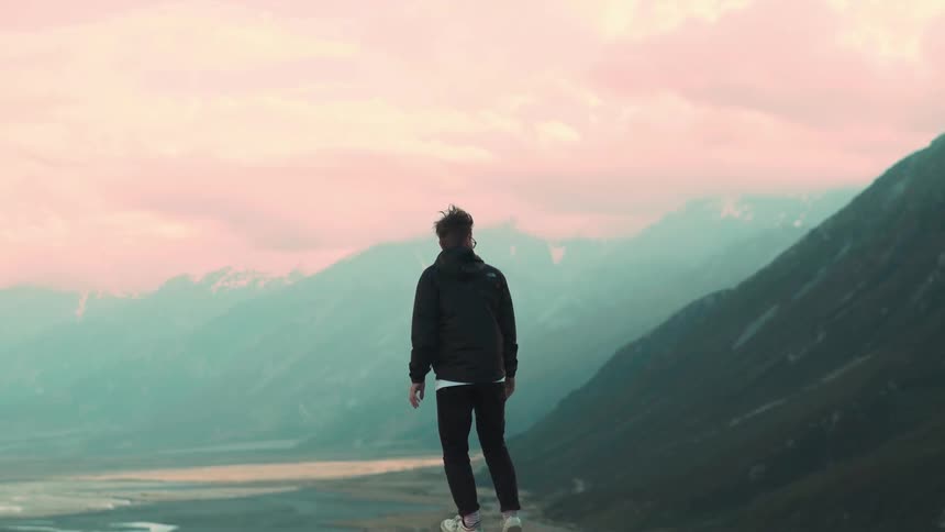 A young man walks to the edge of a viewpoint in slow motion, gazing out at a vast and hazy mountain range under a pink sky