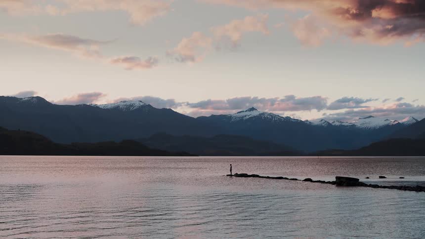A serene, wide shot shows a silhouetted figure standing alone on a rocky pier in a calm lake, looking out at a majestic snow-capped mountain range at sunset