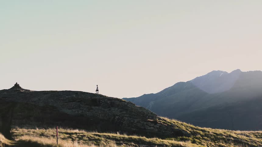 A lone trail runner moves in slow motion along a mountain ridge with a vast alpine landscape in the background during sunrise