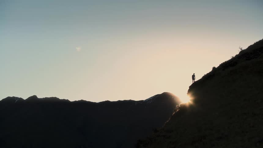 A lone person is silhouetted against the morning sun as they run up a steep and epic mountain ridge