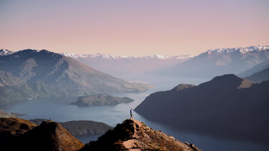 A lone hiker stands atop a mountain peak with arms outstretched, overlooking a vast lake and snow-capped mountain range at sunset